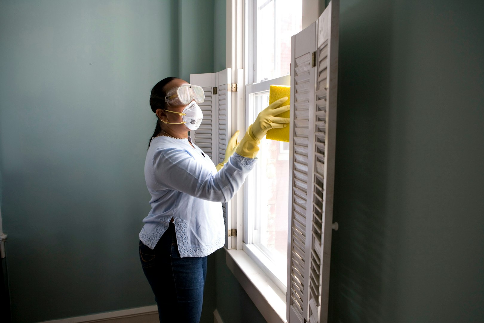 A woman wearing a blue apron and yellow gloves is seen cleaning a workspace filled with plants, natural light, and minimalistic decor. She is focused on organizing items on the table.